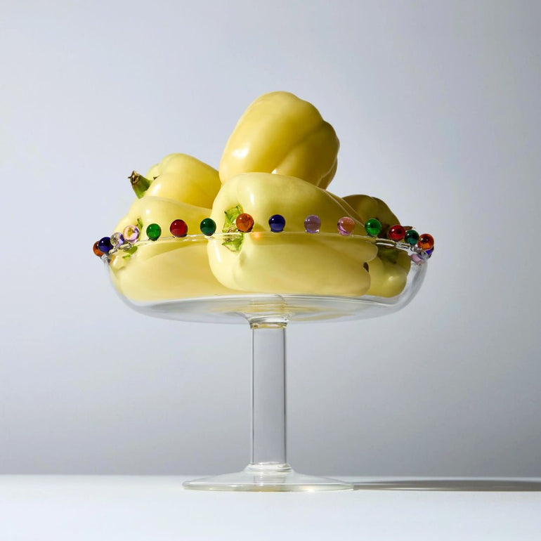 Yellow decorative bell peppers arranged in a clear glass pedestal bowl against a light gray studio background.