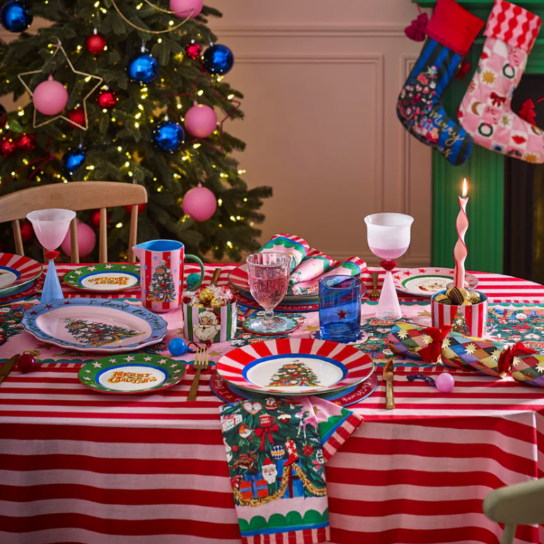 Decorated Christmas table with red and white striped tablecloth, colorful plates, and a Christmas tree in the background.