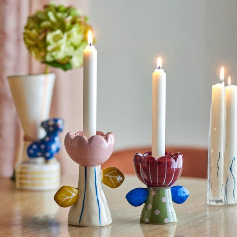 Two decorative ceramic candle holders shaped like stylized flowers with leaf accents, each holding a lit white candle, displayed on a wooden table with other candles and a vase in the softly blurred background.
