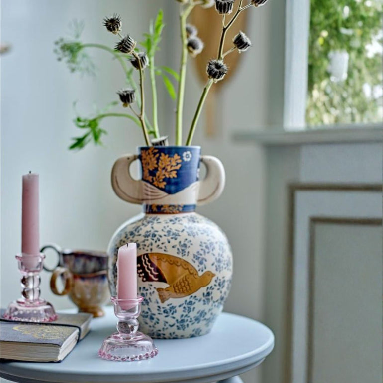 A decorative ceramic vase with a blue and gold floral upper section and a white lower body decorated with blue flowers and a gold bird, holding dried thistle stems on a light blue table. A pink candle in a pink glass holder and another candle in a metal candlestick stand sit beside it.