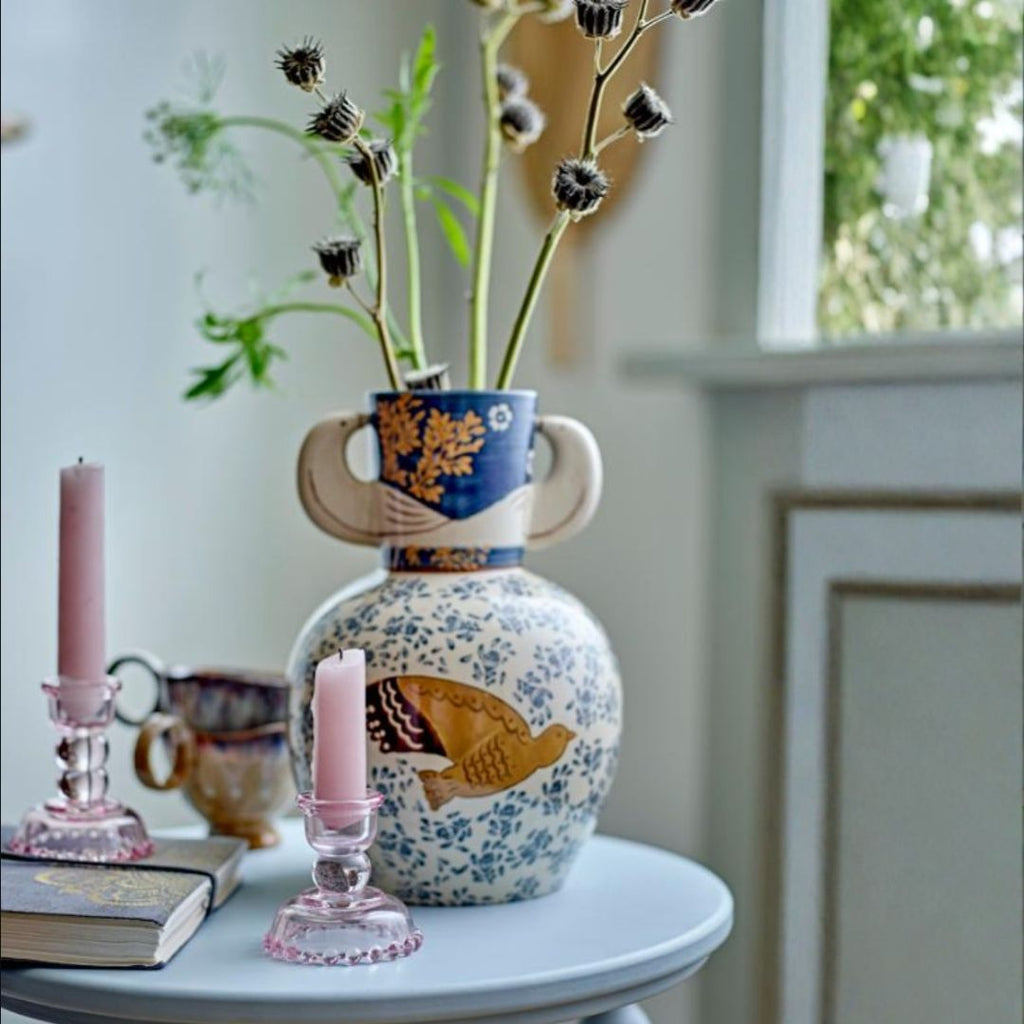 A decorative ceramic vase with a blue and gold floral upper section and a white lower body decorated with blue flowers and a gold bird, holding dried thistle stems on a light blue table. A pink candle in a pink glass holder and another candle in a metal candlestick stand sit beside it.