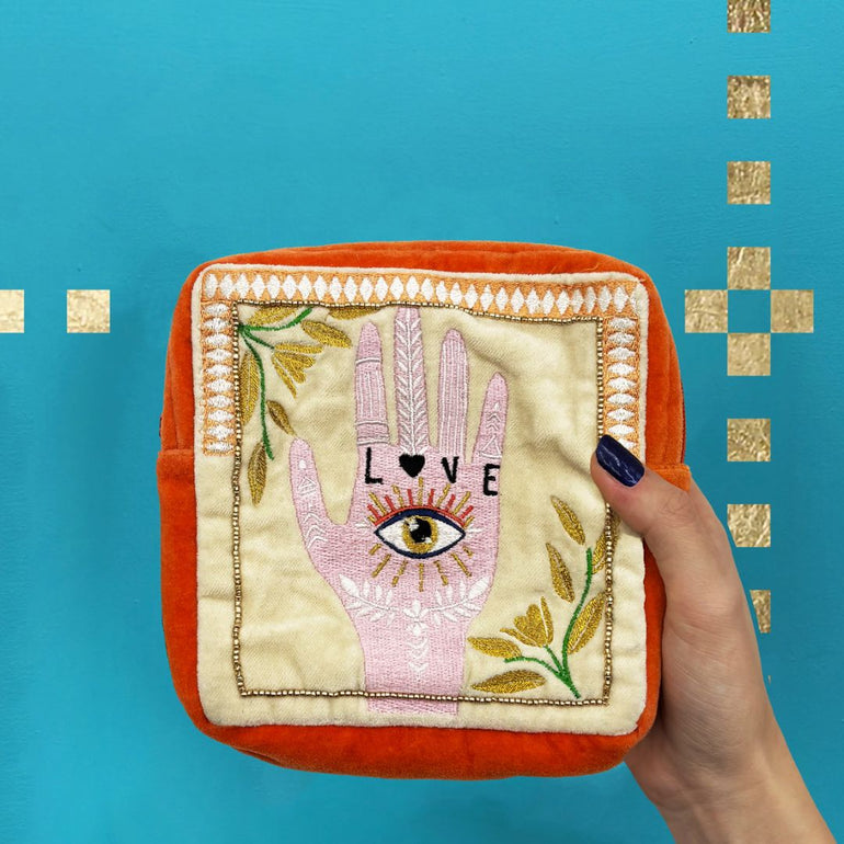 A square orange and cream embroidered pouch featuring a pink Hamsa hand with an eye, gold floral details, and the word 'LOVE' on the front, set against a bright blue background with gold square accents.