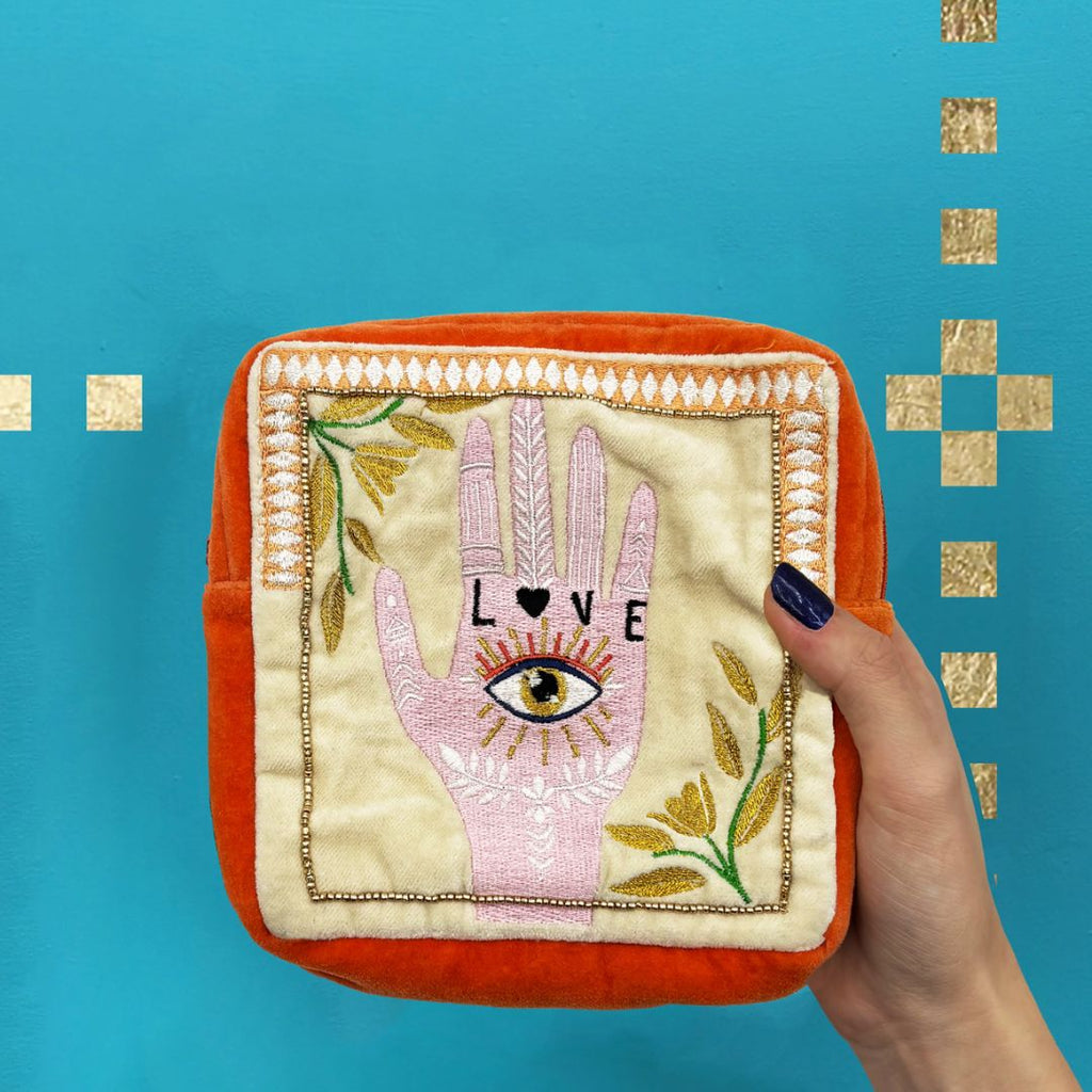 A square orange and cream embroidered pouch featuring a pink Hamsa hand with an eye, gold floral details, and the word 'LOVE' on the front, set against a bright blue background with gold square accents.