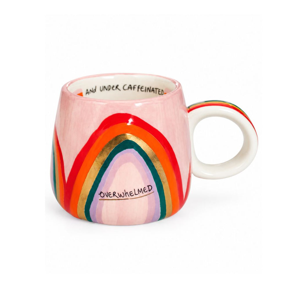 A pink ceramic mug featuring a colorful rainbow design with gold metallic accents and the words 'OVERWHELMED' and 'AND UNDER CAFFEINE' printed on it