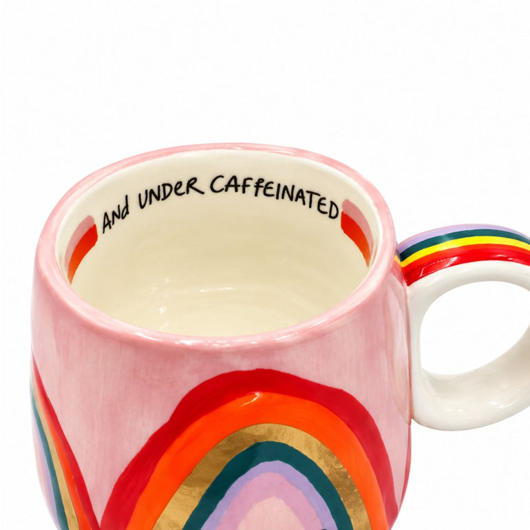 A white ceramic mug with a pink rim, decorated with a colorful rainbow design on the outside and the words 'AND UNDER CAFFEINATED' printed inside.