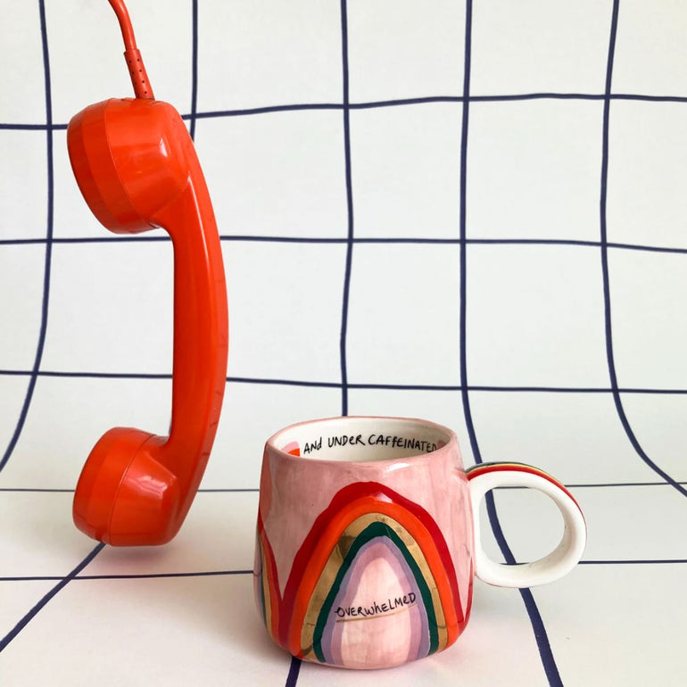 A pink ceramic coffee mug with a colorful hand-painted rainbow design and the word 'OVERWHELMED' on the front, styled next to a bright orange retro telephone receiver against a white background with a dark blue grid pattern.