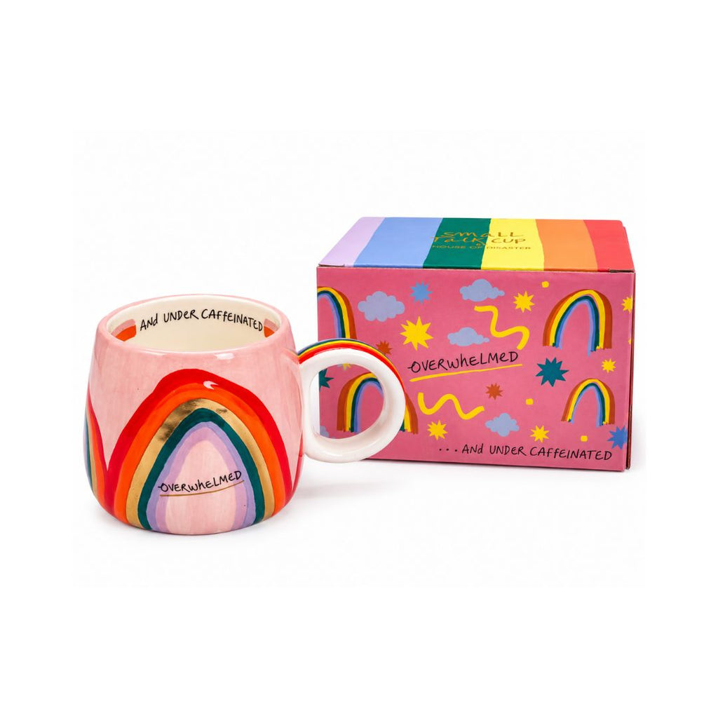 A colorful ceramic mug with a rainbow design next to its matching pink and rainbow gift box, featuring the text 'AND UNDER CAFFEINIZED' and 'OVERWHELMED'.