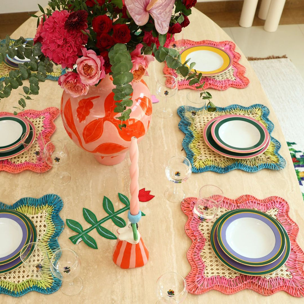 A styled dining table setting featuring colorful crocheted placemats, stacked white plates with striped rims, a floral vase filled with pink and red flowers, and a candle on a light wood table.