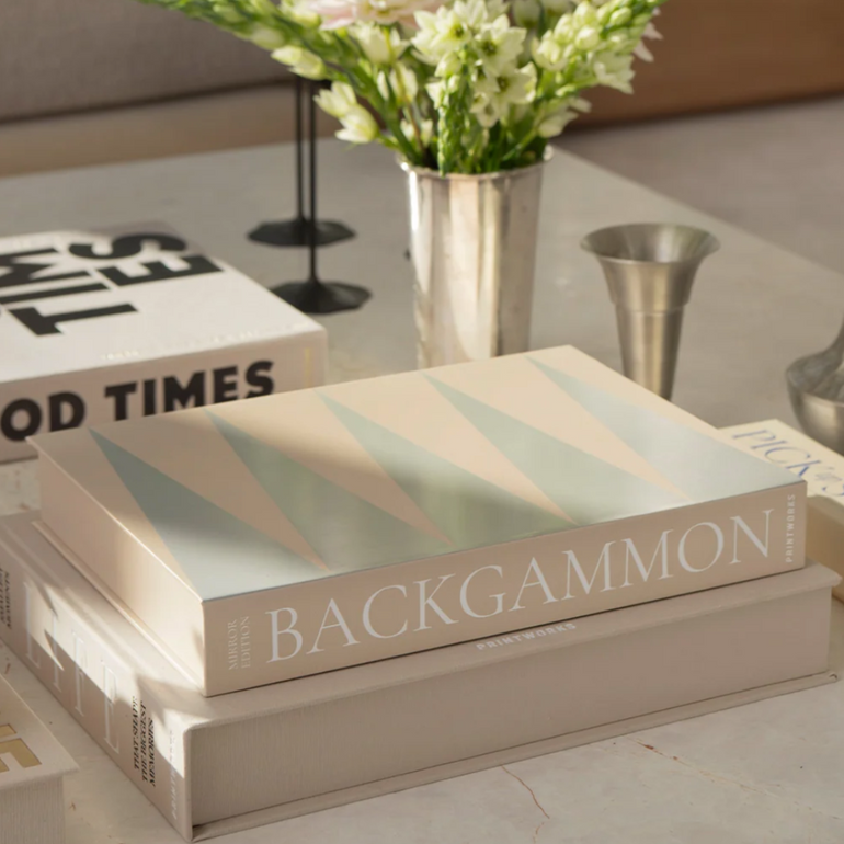 A stack of three decorative books on a light marble table, with the top book titled BACKGAMMON. The setting includes a vase of white flowers, metal candlesticks, and other styled books in a softly lit interior.